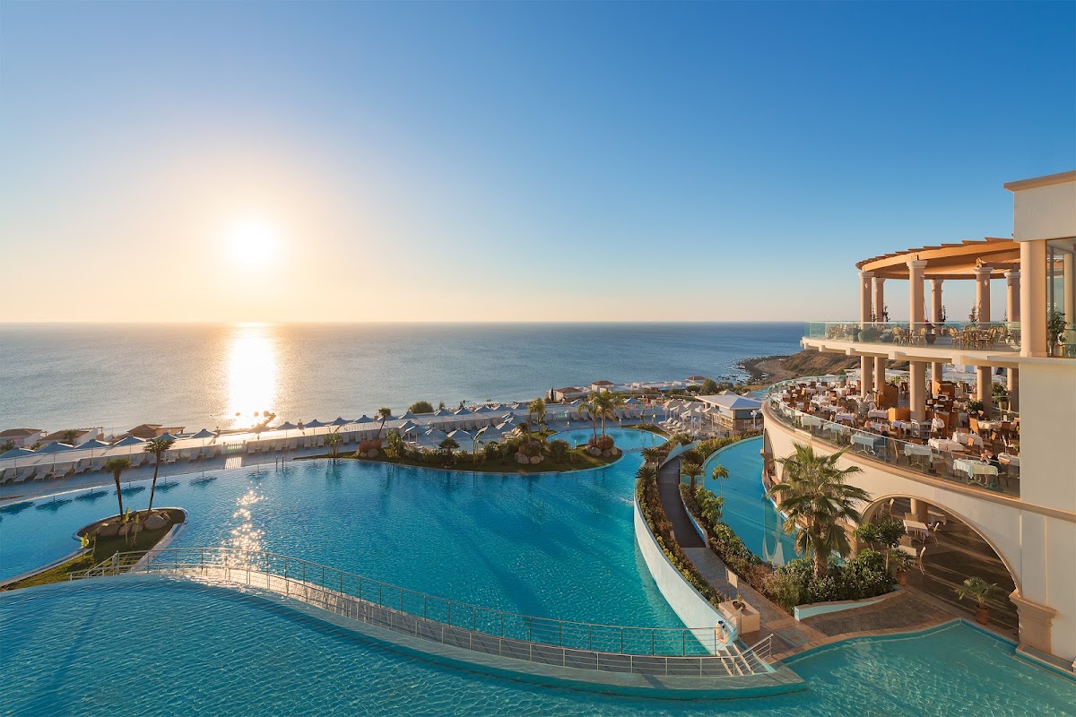 Atrium Prestige Thalasso Spa Resort overlooks curved pools with palm trees and a Mediterranean coastline at sunset.