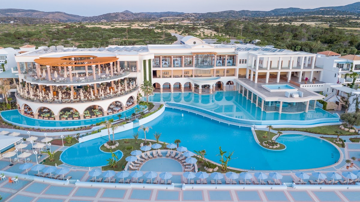 Atrium Prestige Thalasso Spa Resort features curved pools, white buildings with orange accents, and loungers arranged around landscaped islands, backed by green hills.