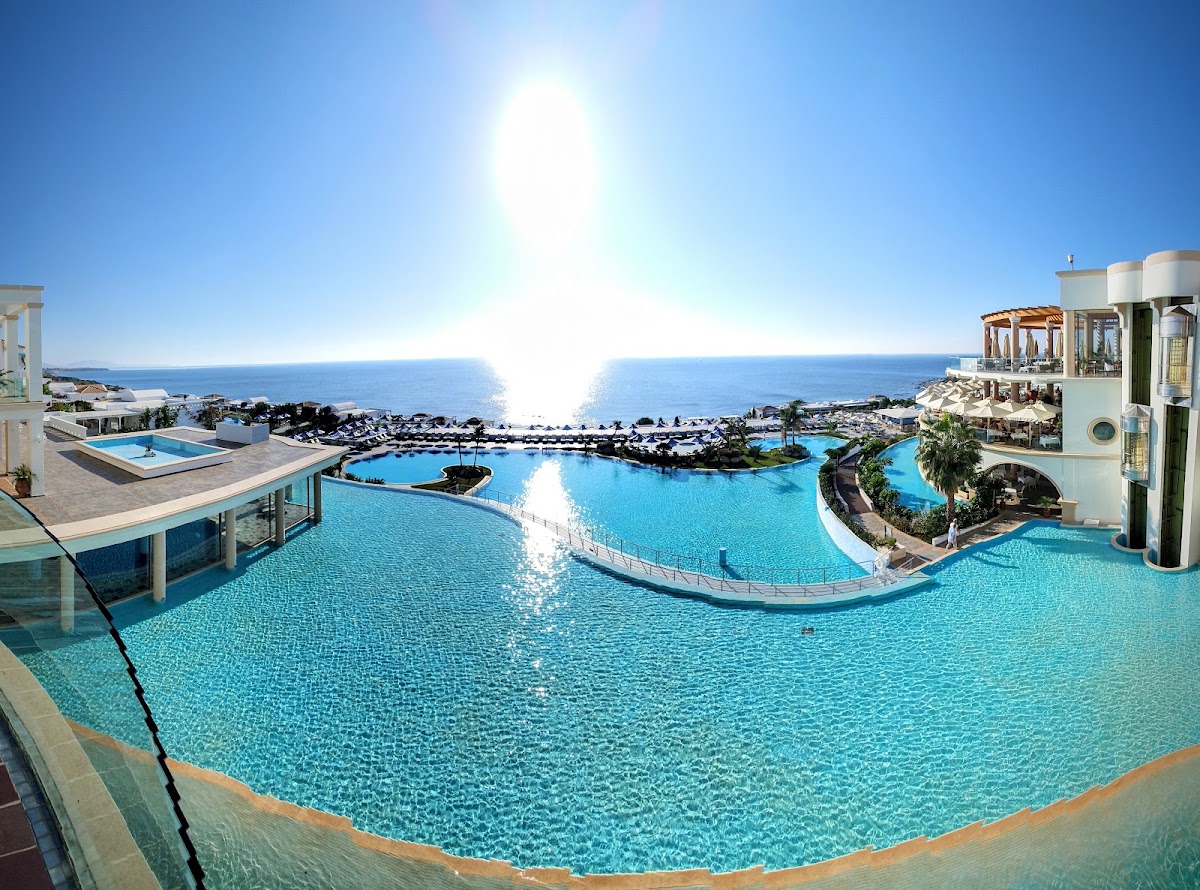 Curved turquoise pool with islands and lounging areas overlooking the Mediterranean Sea at Atrium Prestige Thalasso Spa Resort and Villas under bright sunlight.