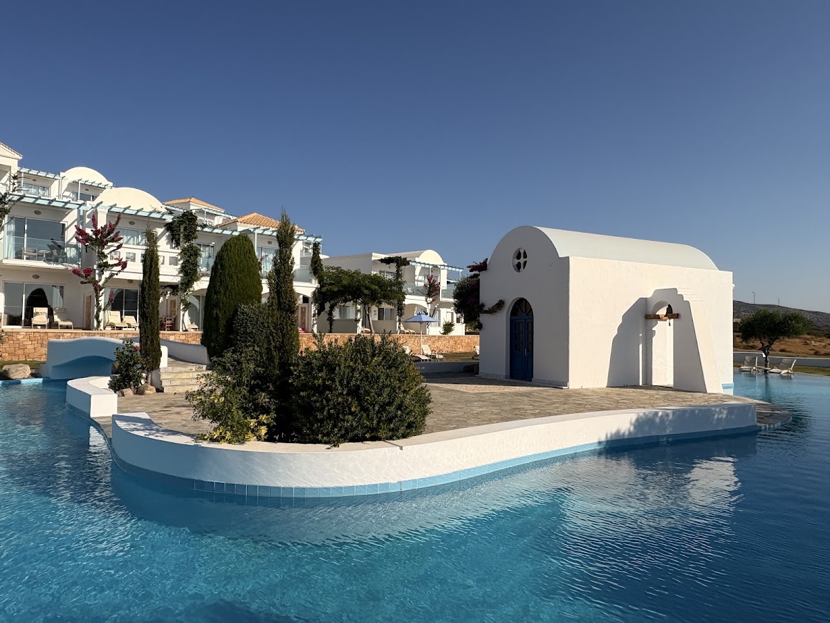 White chapel with blue door surrounded by pool water, cypress trees, and resort buildings under clear sky at Atrium Prestige Thalasso Spa Resort.