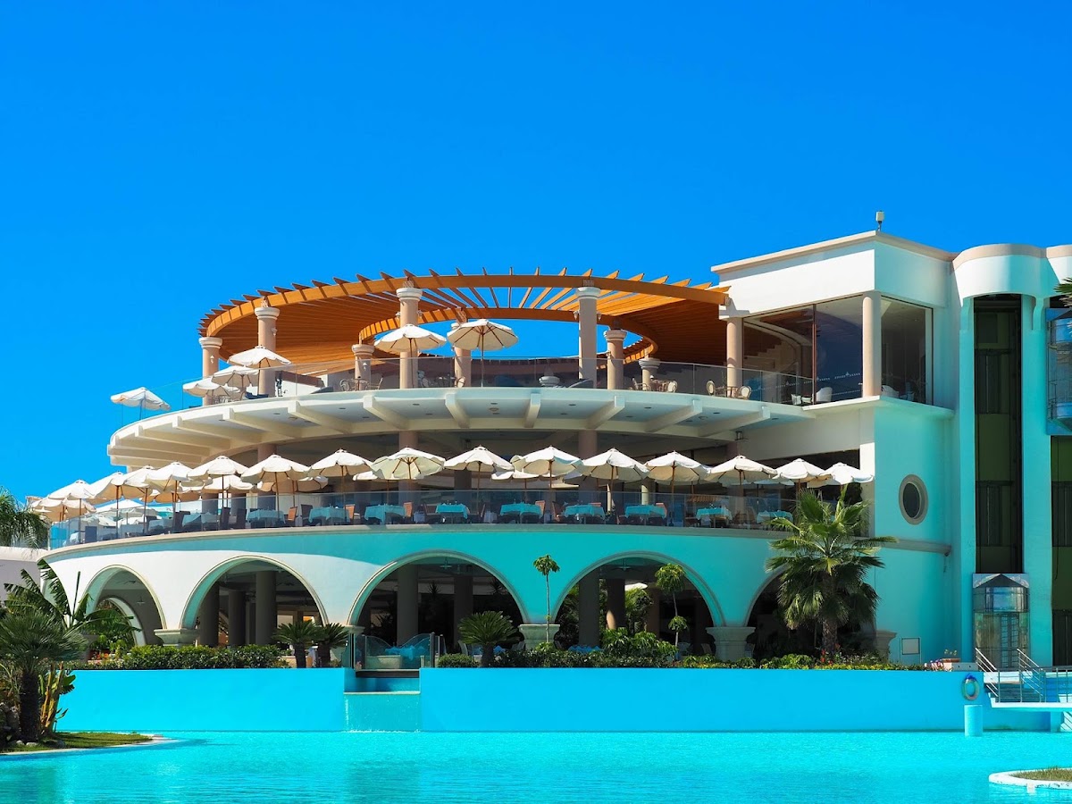 Turquoise pool fronts a modern white and aqua resort building with arched openings, shaded seating areas, and wooden pergolas under clear blue sky.
