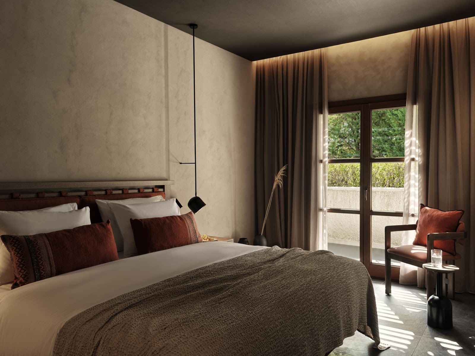 Modern resort bedroom with bed dressed in gray linens and burgundy pillows, pendant light, beige walls, and French doors opening to a garden terrace.