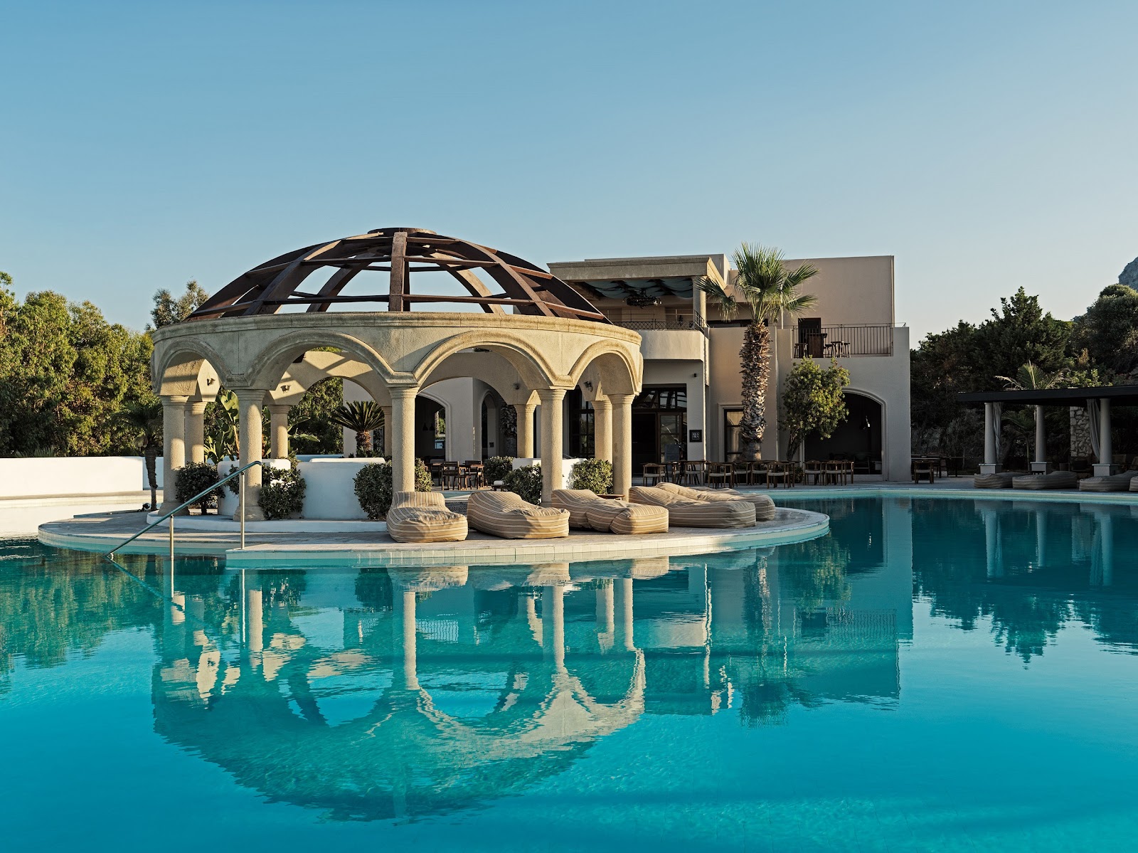 Turquoise pool surrounding a domed pavilion with arched columns at Aulūs Lindos Rhodes Resort, with white resort buildings and palm trees behind.