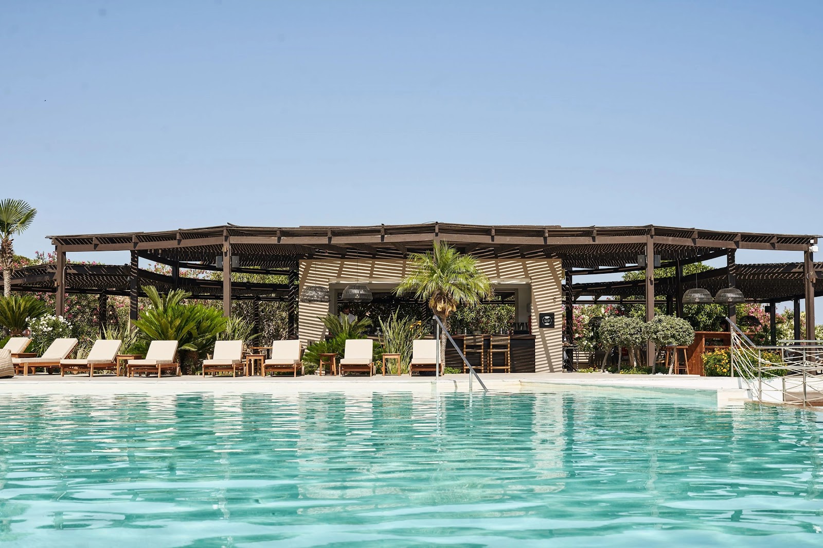 Swimming pool with turquoise water facing a wooden pergola structure with lounge chairs, palm trees, and a bar pavilion at Aulūs Lindos Rhodes Resort.
