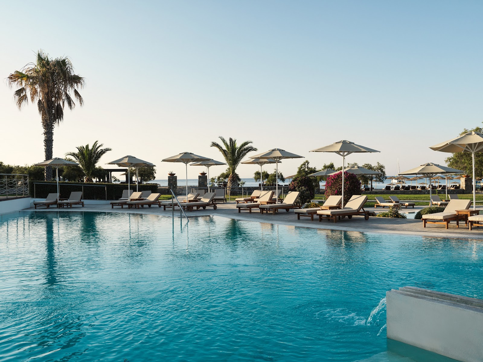 Large resort pool with lounge chairs and umbrellas, palm trees, and sea view in the background under clear sky.