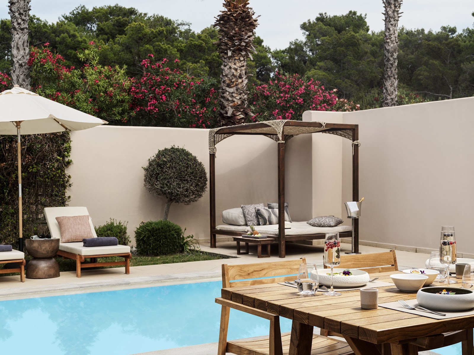Pool terrace with wooden dining table, cabana bed, loungers, white umbrellas, and magenta bougainvillea beyond cream walls at Aulūs Lindos Rhodes Resort.