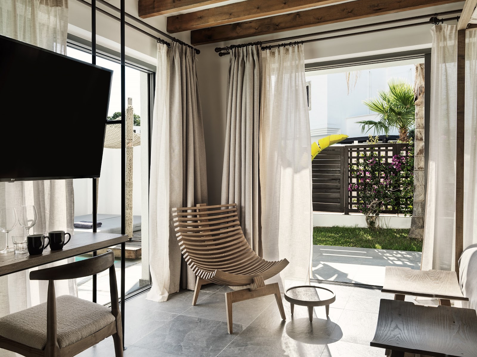 Modern resort suite with wooden beams, TV, striped lounge chair, and open doors leading to a patio with palm trees and ocean view.