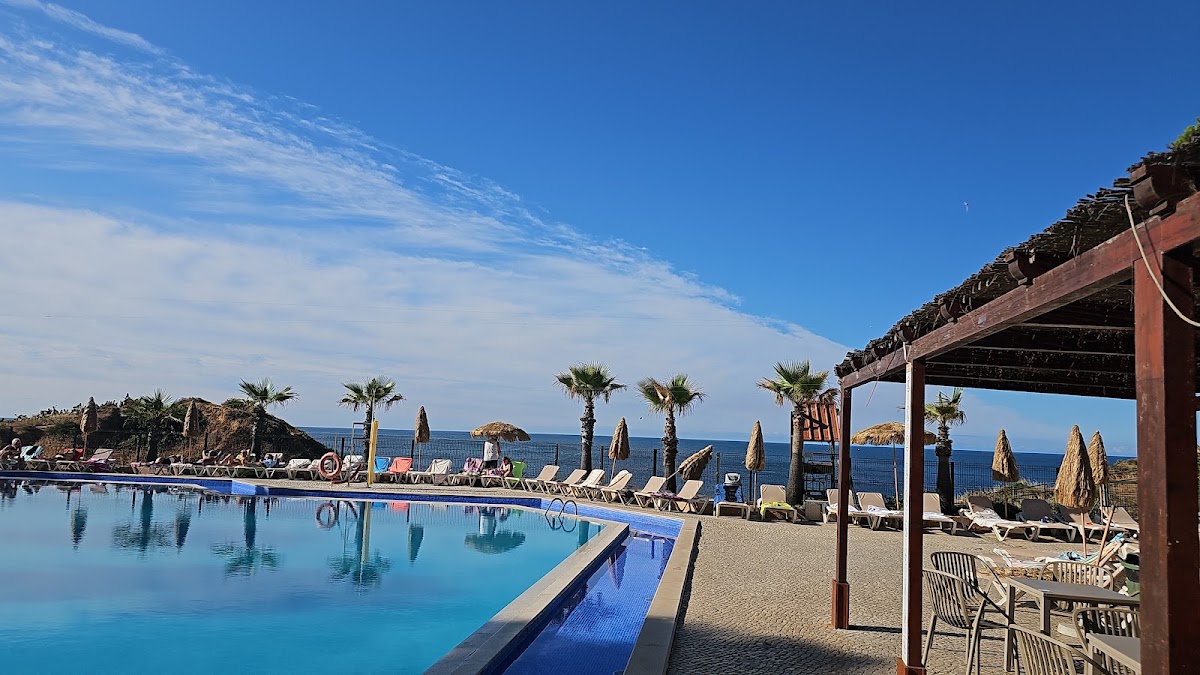 Auramar's clifftop pool overlooks the Mediterranean Sea with lounge chairs, umbrellas, and palm trees lining the beach below.