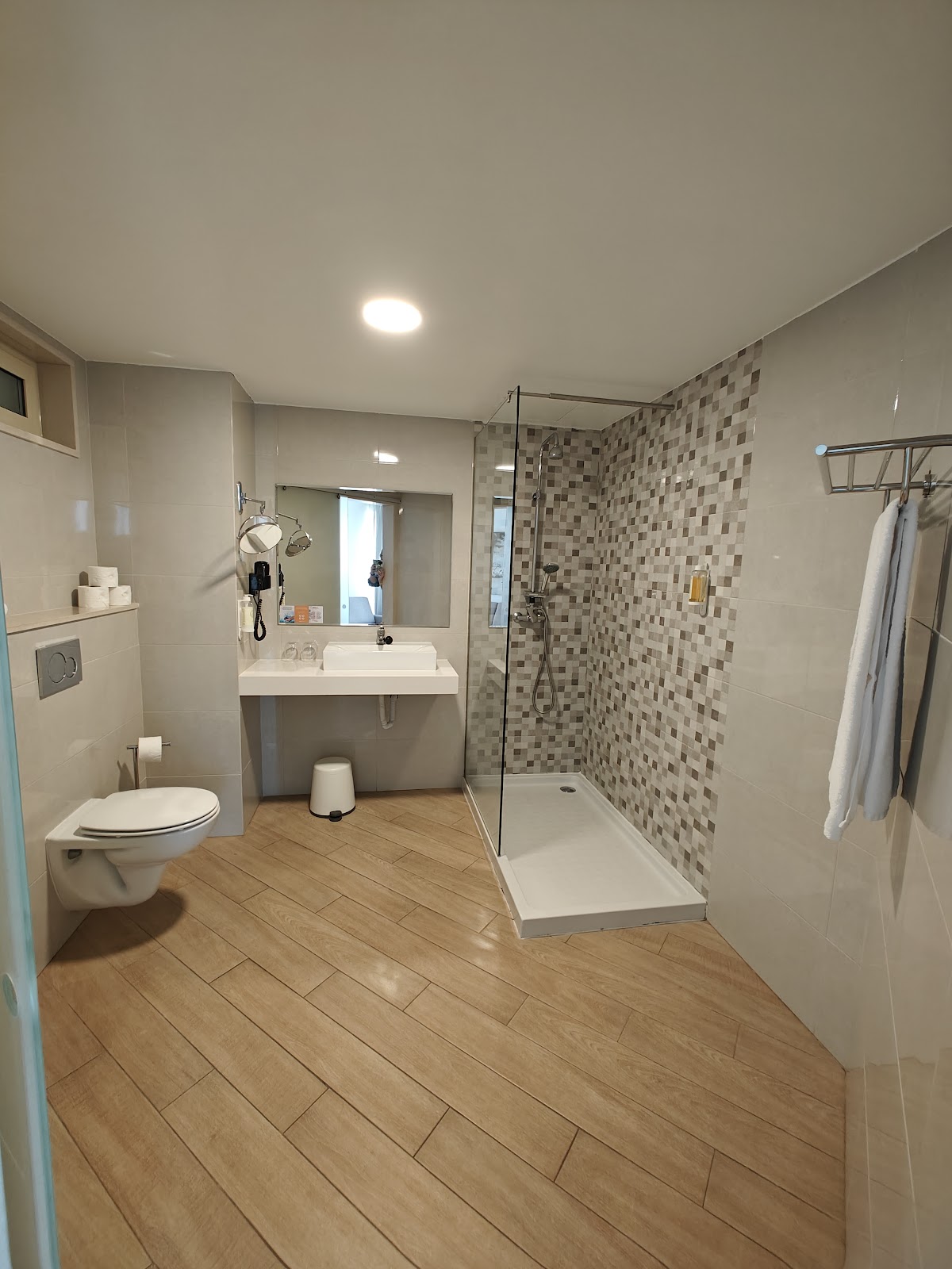 Modern bathroom with wood-look flooring, white sink vanity with mirror, walk-in shower with mosaic tile surround, and toilet at Auramar resort.