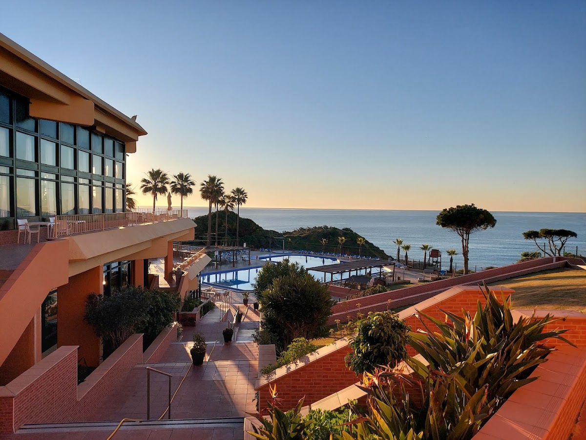 Terracotta resort buildings overlook a curved pool and Mediterranean Sea at sunset, with palm trees and tropical plants dotting the clifftop grounds.