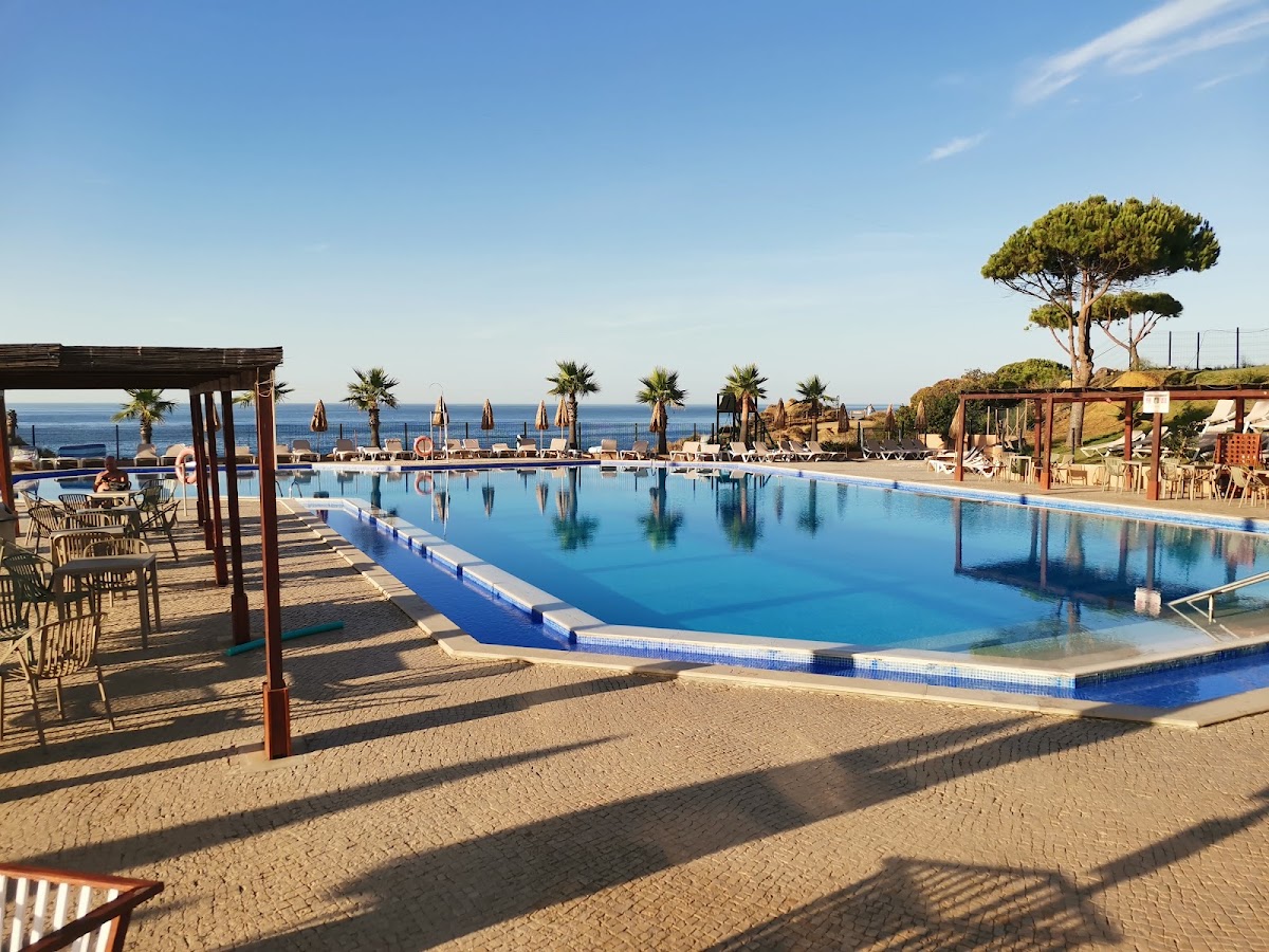 Large resort pool with blue water overlooking the Mediterranean Sea, lined with palm trees and beach loungers under pergolas.