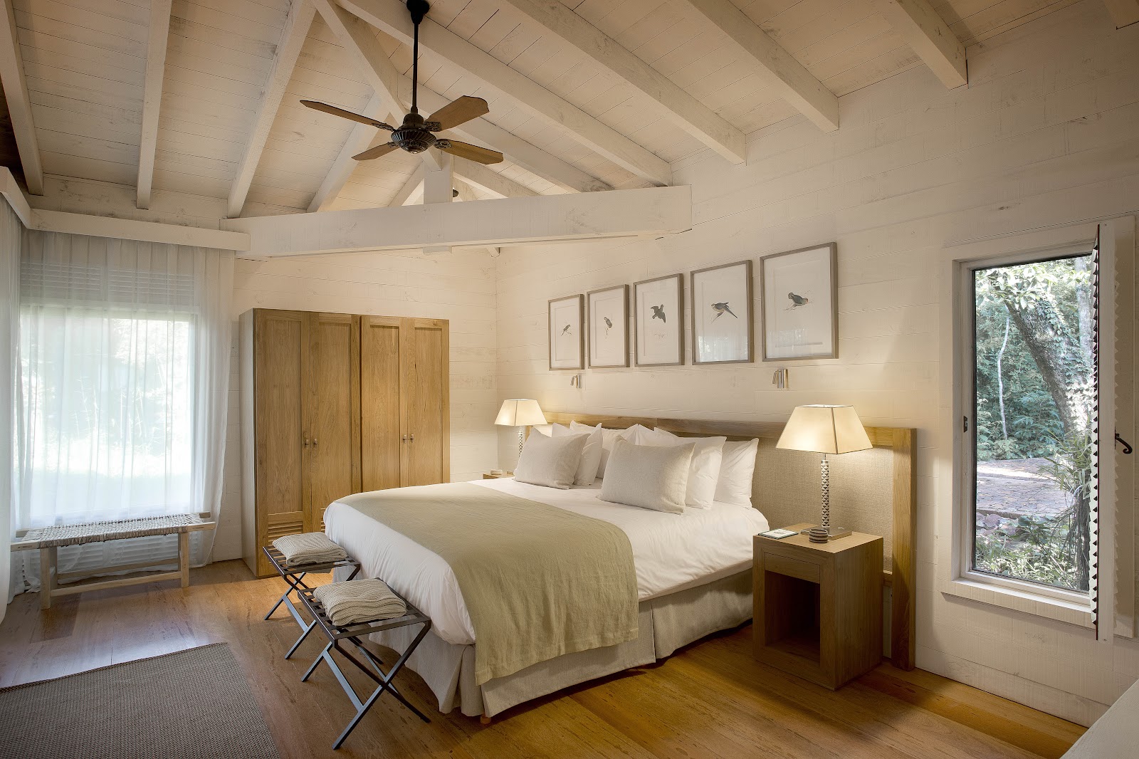 Bright bedroom with vaulted white ceiling, ceiling fan, king bed with cream bedding, wooden nightstands with lamps, and glass doors opening to forest views.