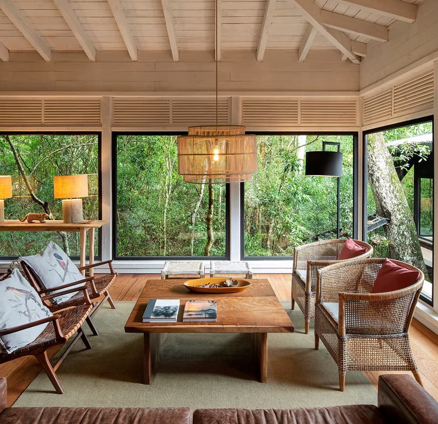 Modern lounge with wooden furniture and woven chairs overlooks lush forest through floor-to-ceiling windows at Awasi Iguazú.
