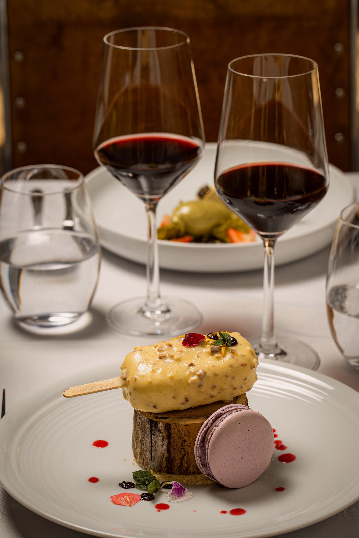 Fine dining dessert plate with yellow mousse popsicle, chocolate macaron, and garnishes alongside red wine glasses and water at Awasi Mendoza.