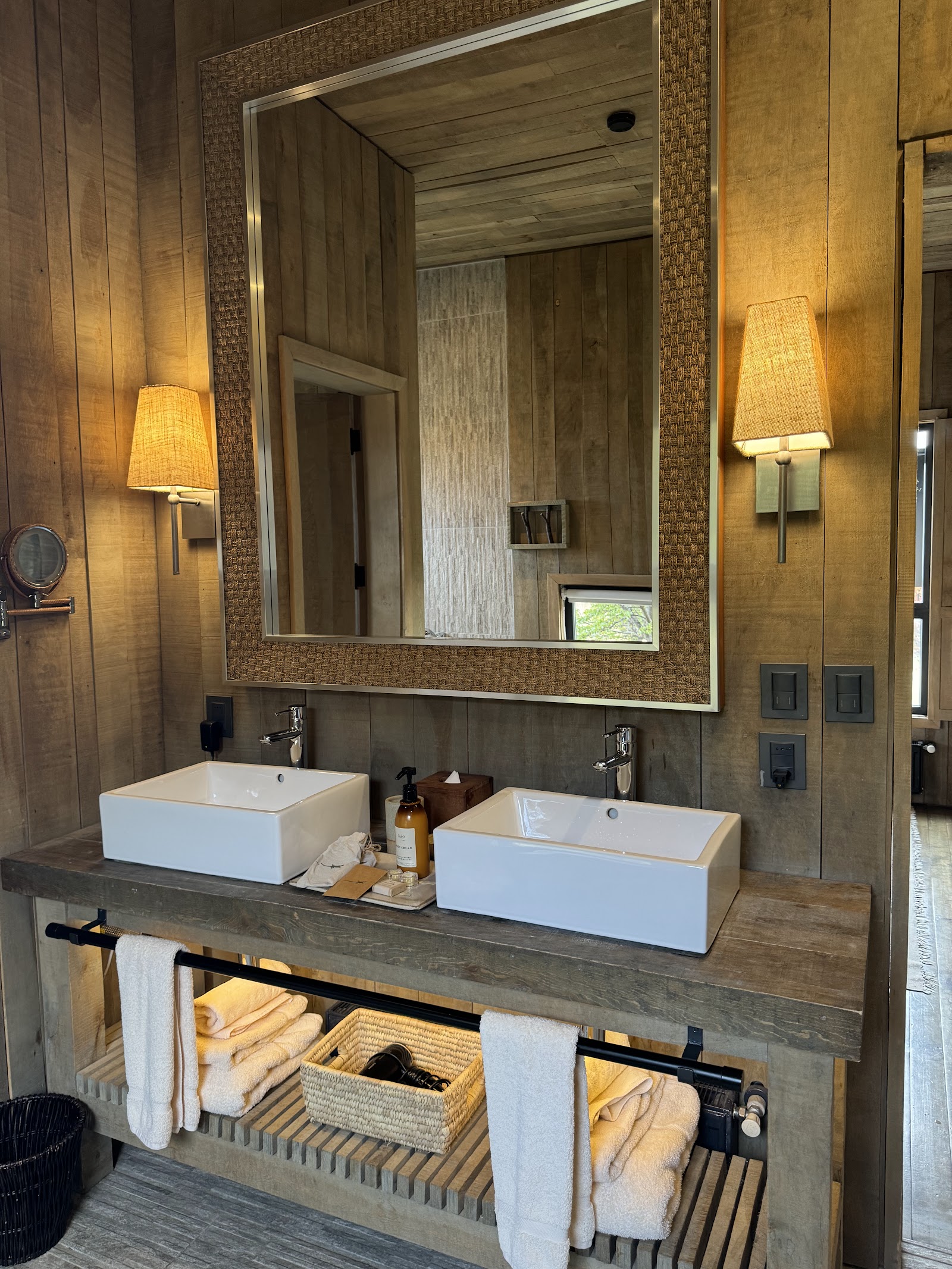 Resort bathroom with dual white vessel sinks, large framed mirror, warm wall sconces, and wooden vanity with woven storage baskets.