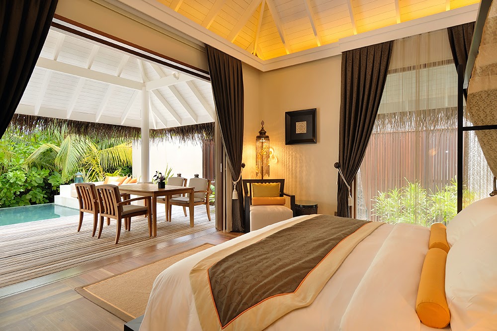 Tropical overwater villa interior with bed, dining area, and private pool visible through open doors under thatched roof.