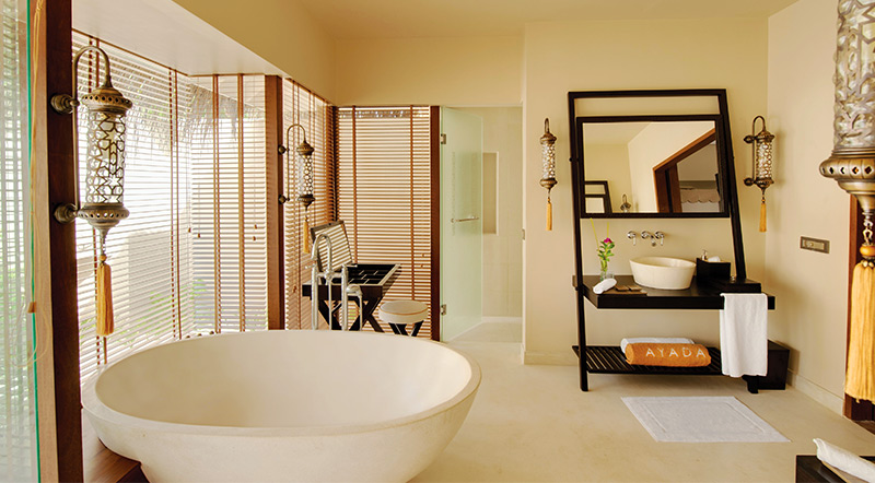 Spa-style bathroom with a freestanding round soaking tub, wooden room divider screen, and decorative mirror with black frame at Ayada Maldives.