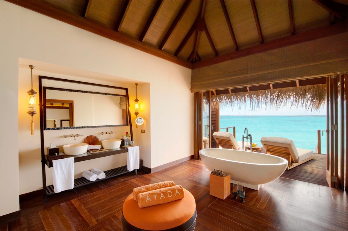 Overwater villa bathroom at Ayada Maldives with wooden beamed ceiling, double vanity, freestanding tub, and ocean view through open doors to deck with loungers.