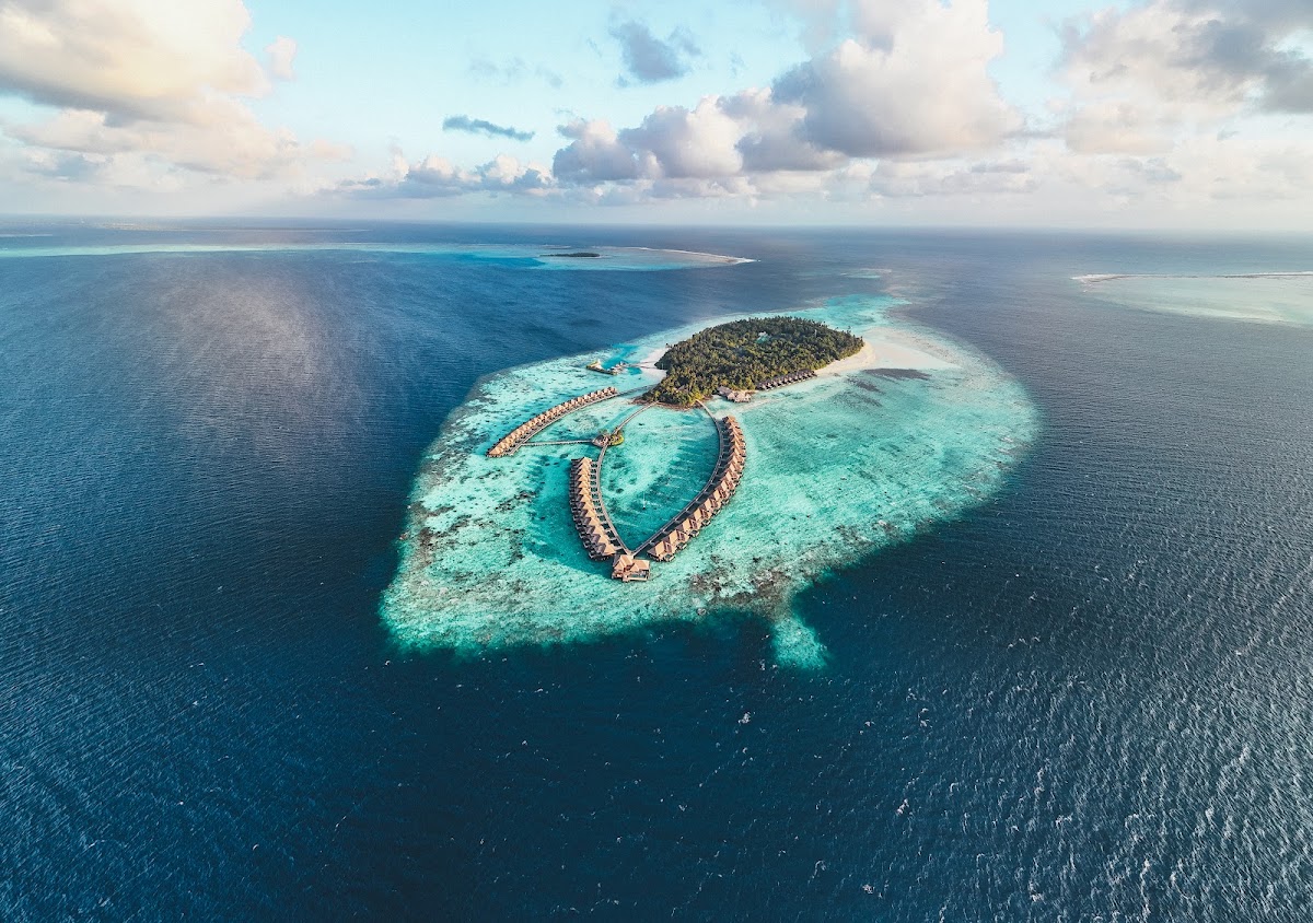 Aerial view of Ayada Maldives island surrounded by turquoise lagoon and coral reef, with overwater bungalows arranged in curved rows.