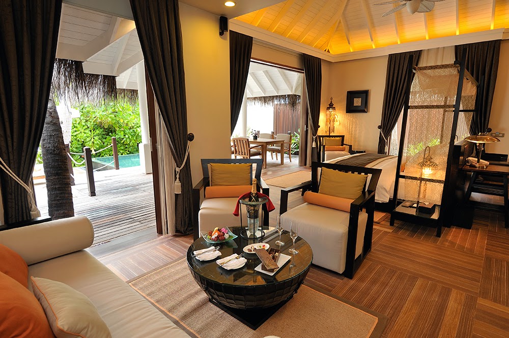 Spacious villa interior at Ayada Maldives with cream and orange furnishings, wooden floors, and open doors revealing a pool and tropical garden beyond.