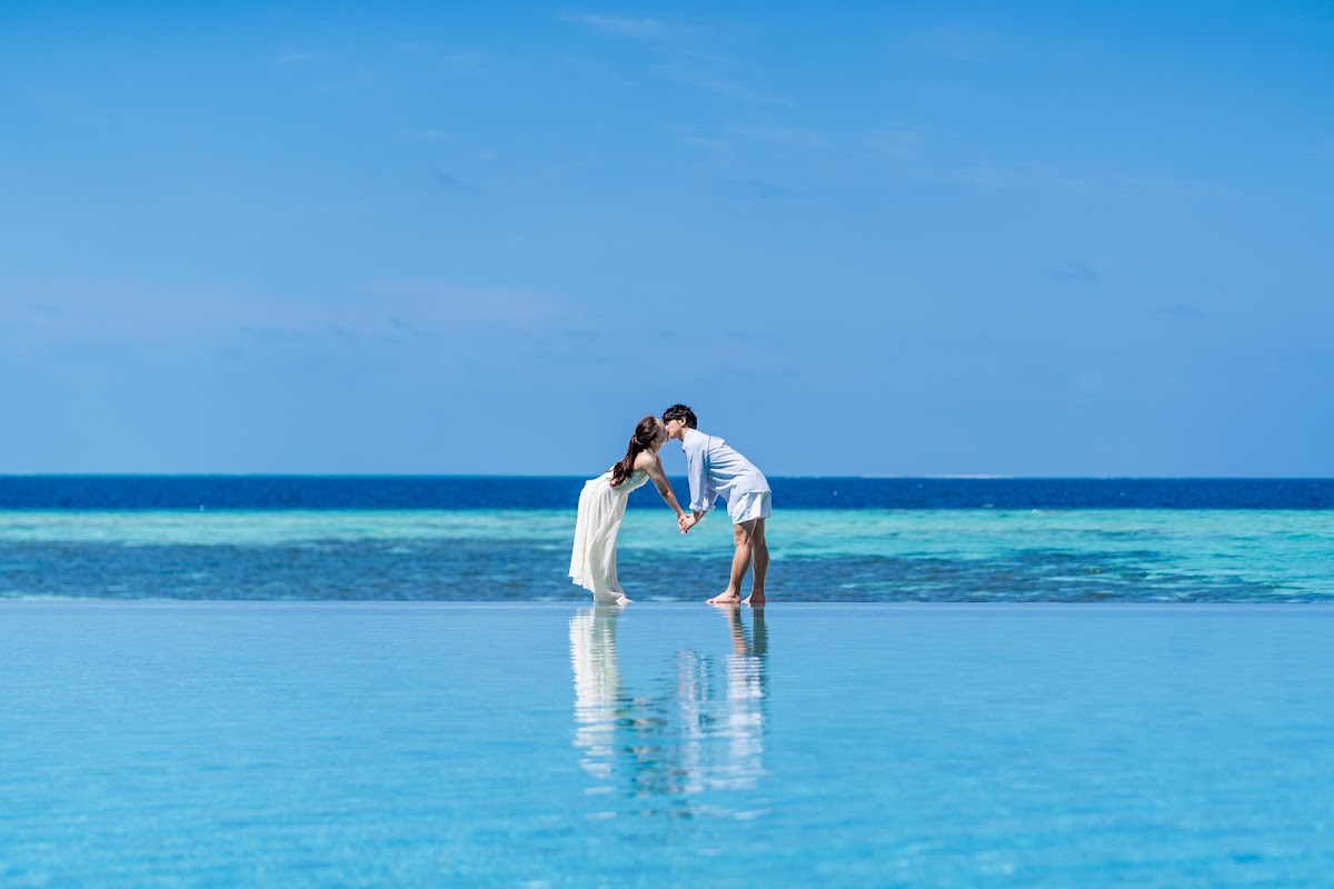Couple in white clothing kiss in shallow turquoise water with calm ocean and clear blue sky at Ayada Maldives.