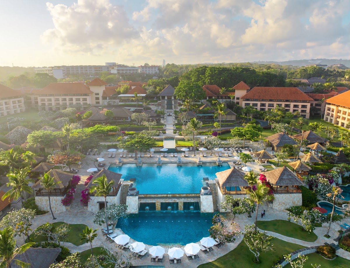 AYANA Resort Bali's twin lagoon pools surrounded by loungers, thatched-roof structures, manicured gardens, and multi-story accommodations amid lush tropical landscape.