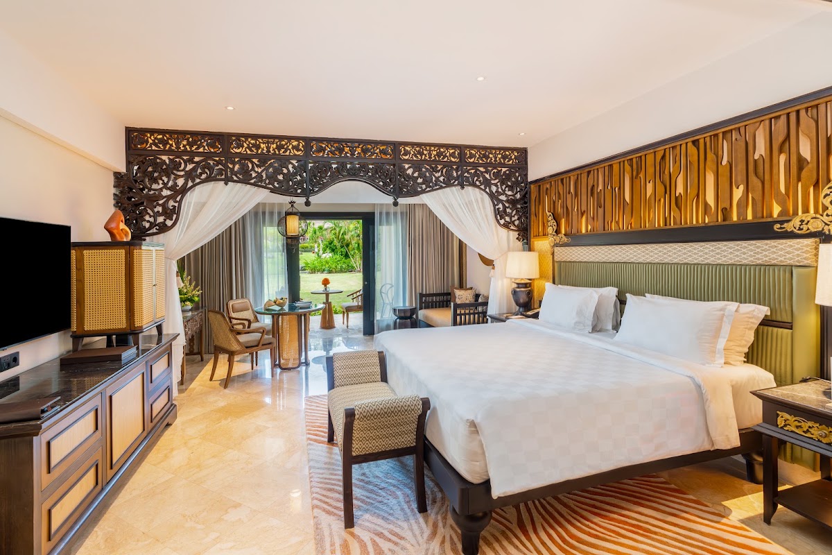 Spacious resort bedroom at AYANA with two queen beds, ornate carved headboard, and garden view through glass doors.