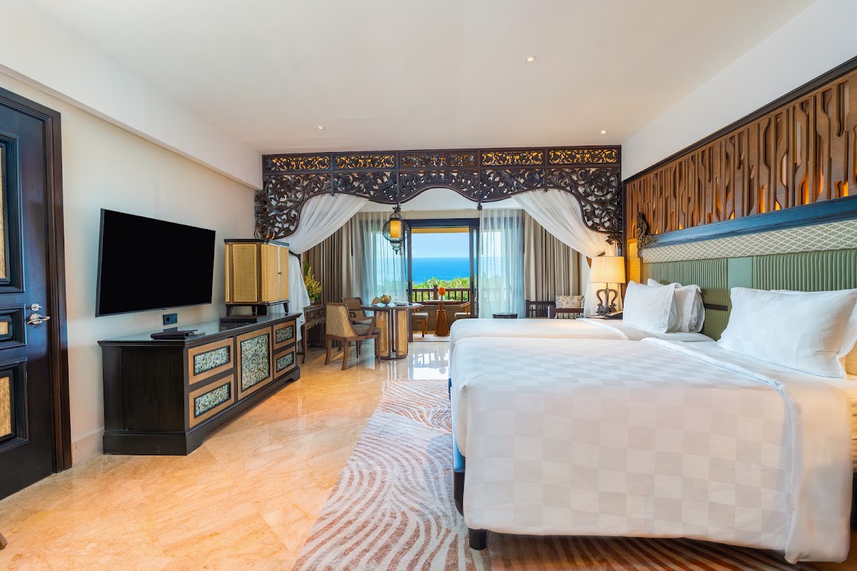 Spacious resort suite with two beds, ornate carved headboard, ocean view balcony, and flat-screen TV on dark wood console.
