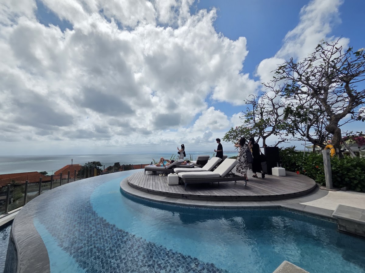 Curved infinity pool with loungers on a wooden deck overlooking the ocean at AYANA Resort Bali, with guests and a gnarled tree visible.
