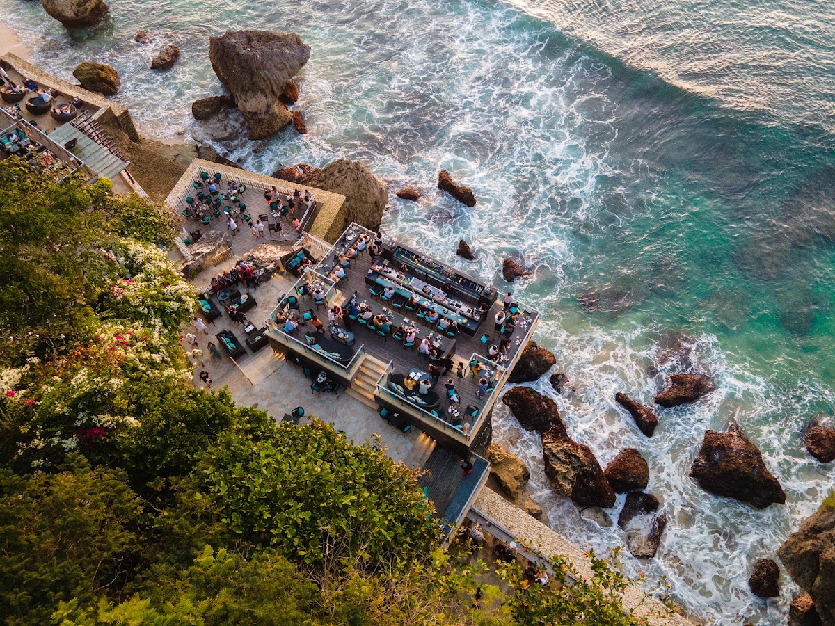 Aerial view of AYANA Resort Bali's clifftop terrace with guests dining and lounging above turquoise waves and rocky outcrops.