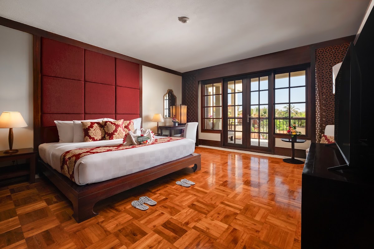 Spacious resort bedroom with red upholstered headboard, wooden platform bed, and French doors opening to a balcony overlooking tropical gardens.