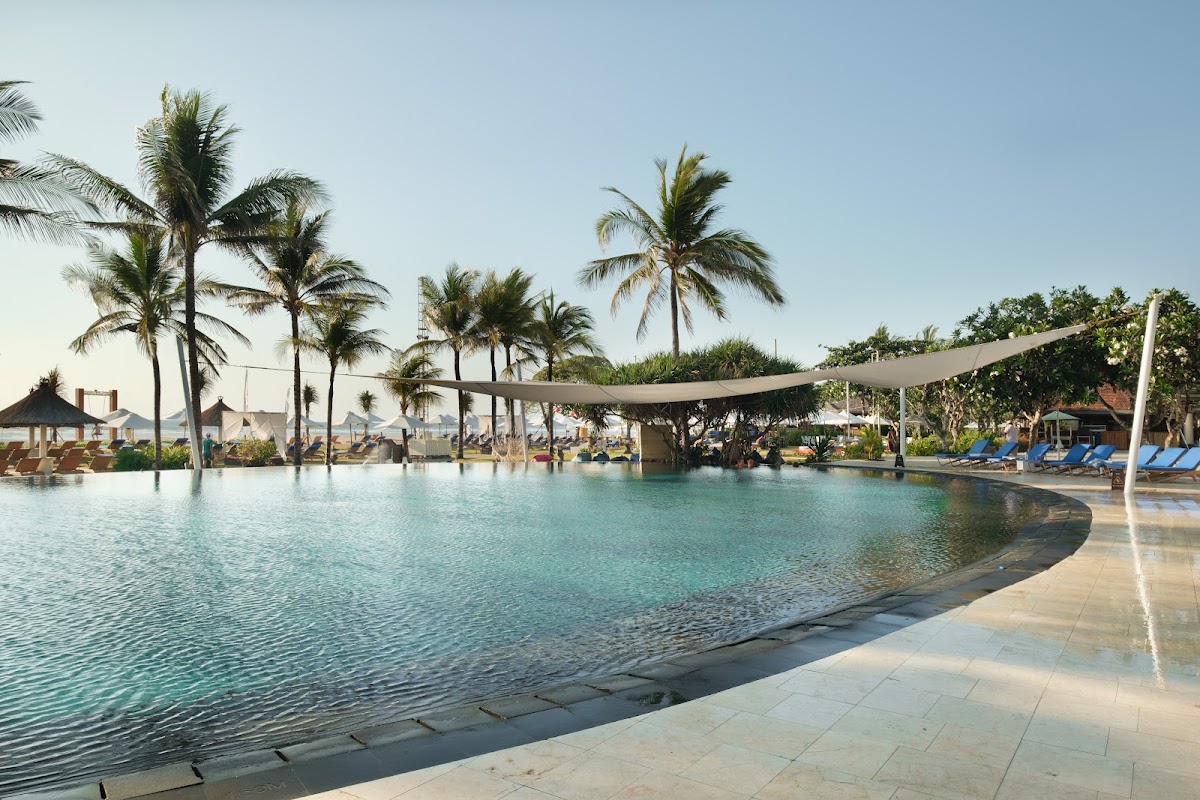 Lagoon-style pool at Ayodya Resort Bali with palm trees, sun loungers, and thatched umbrellas under clear sky.