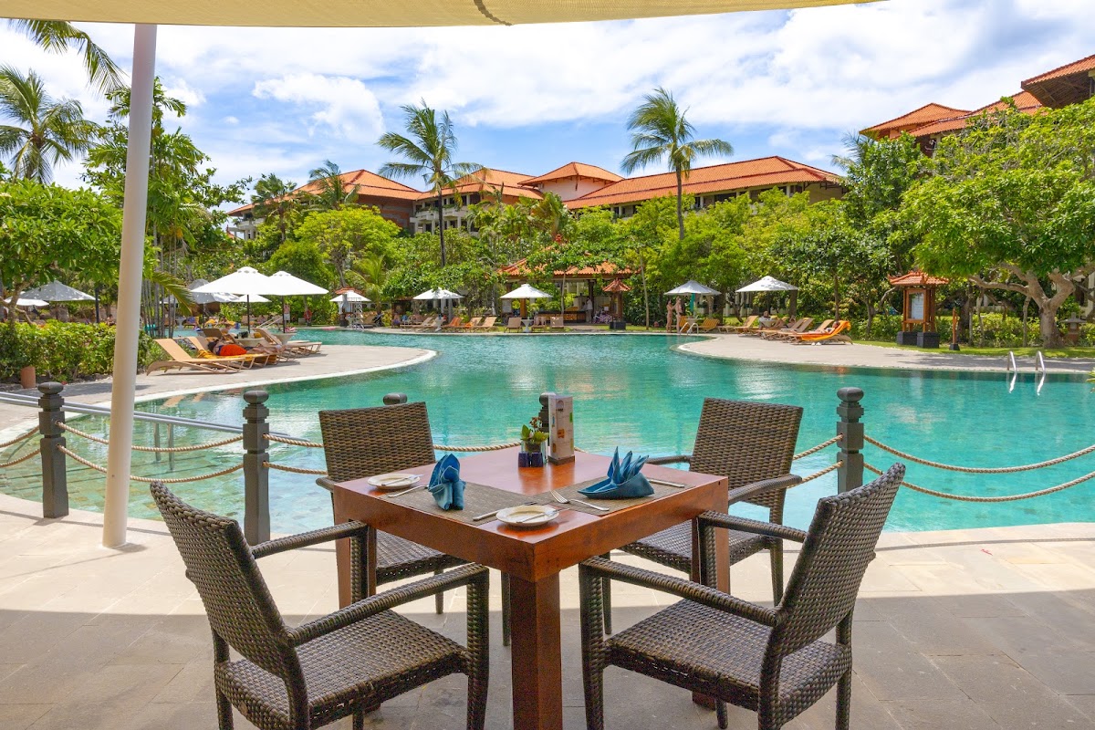 Dining table with woven chairs under a tan shade sail overlooks a turquoise pool surrounded by white umbrellas, loungers, and tropical landscaping at Ayodya Resort Bali.
