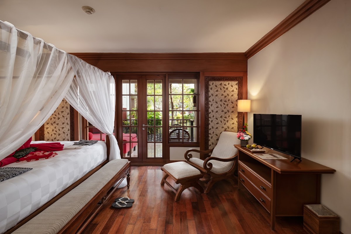 Resort suite with four-poster canopy bed, wooden furnishings, and glass doors opening to a tropical garden balcony.