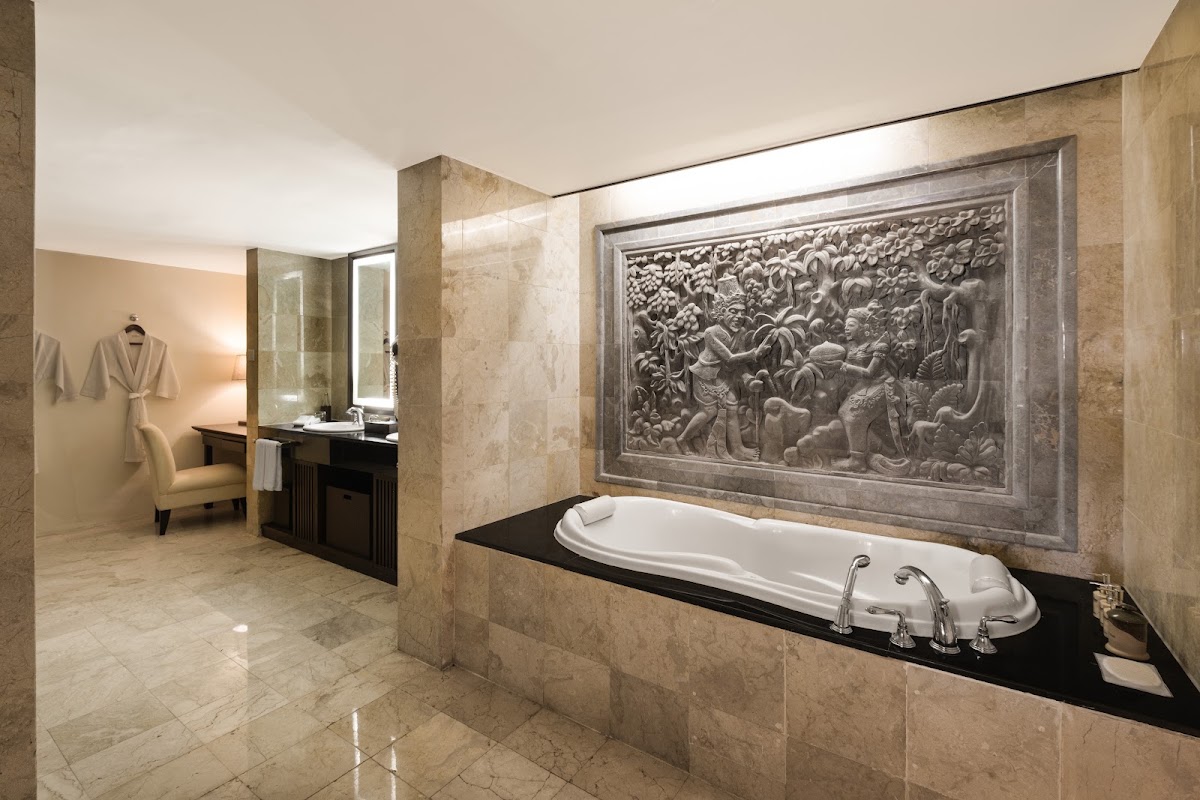 Bathroom suite with sunken white bathtub, ornate stone relief panel above, marble flooring and walls, and vanity area with chair at Ayodya Resort Bali.
