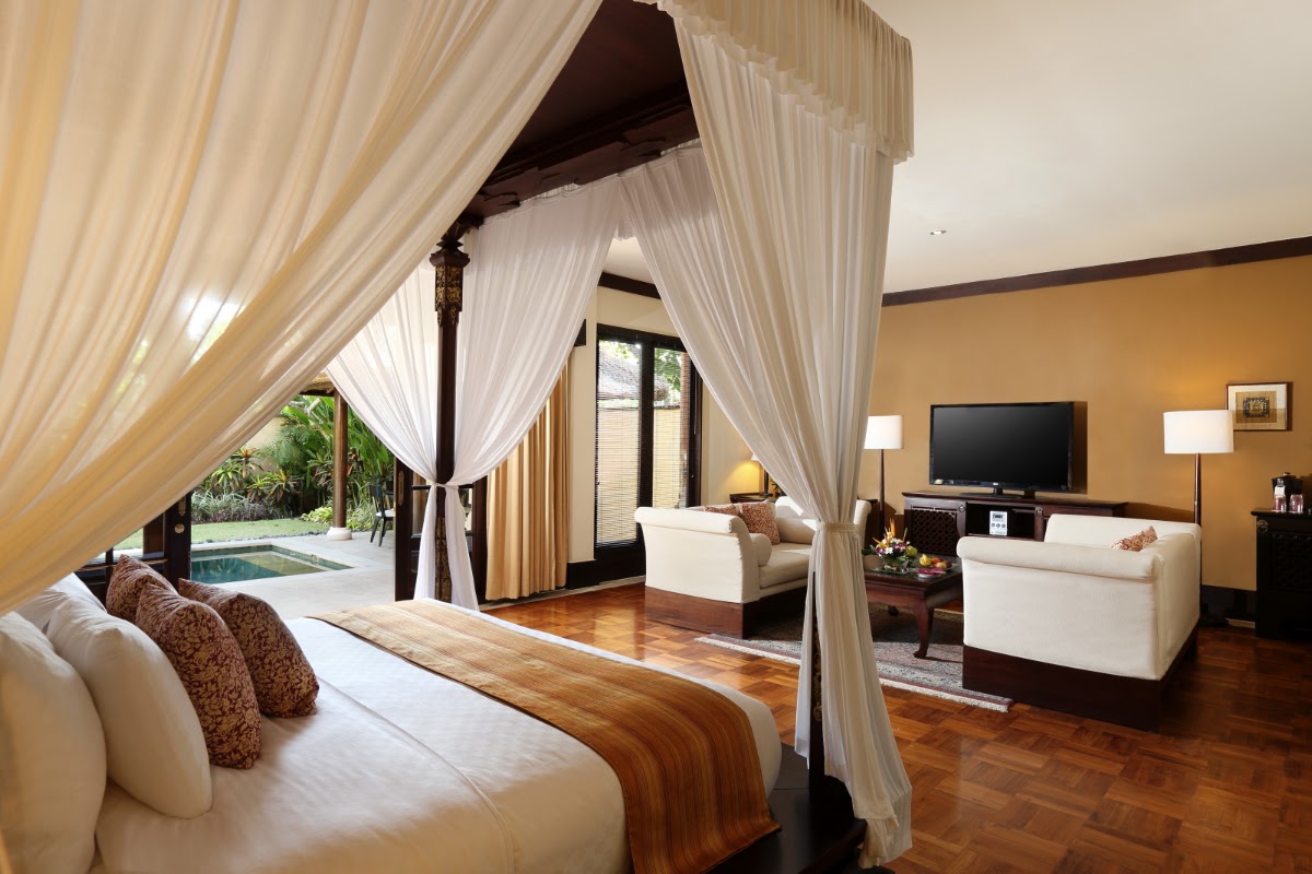 Canopied bed with cream drapes opens to a spacious suite with wood floors, sitting area, and view of private pool and garden at Ayodya Resort Bali.