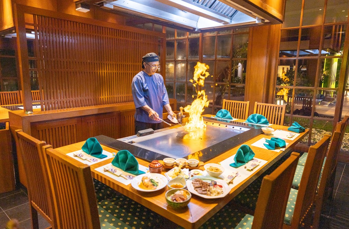 Chef prepares teppanyaki at a wooden counter with flames while diners sit around the table with plated dishes and green napkins.