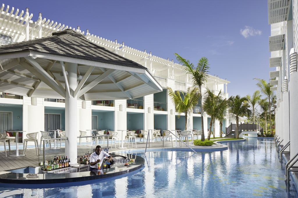 Swim-up bar with bartender serving drinks in resort pool surrounded by white cabanas, palm trees, and high-rise buildings under clear sky.