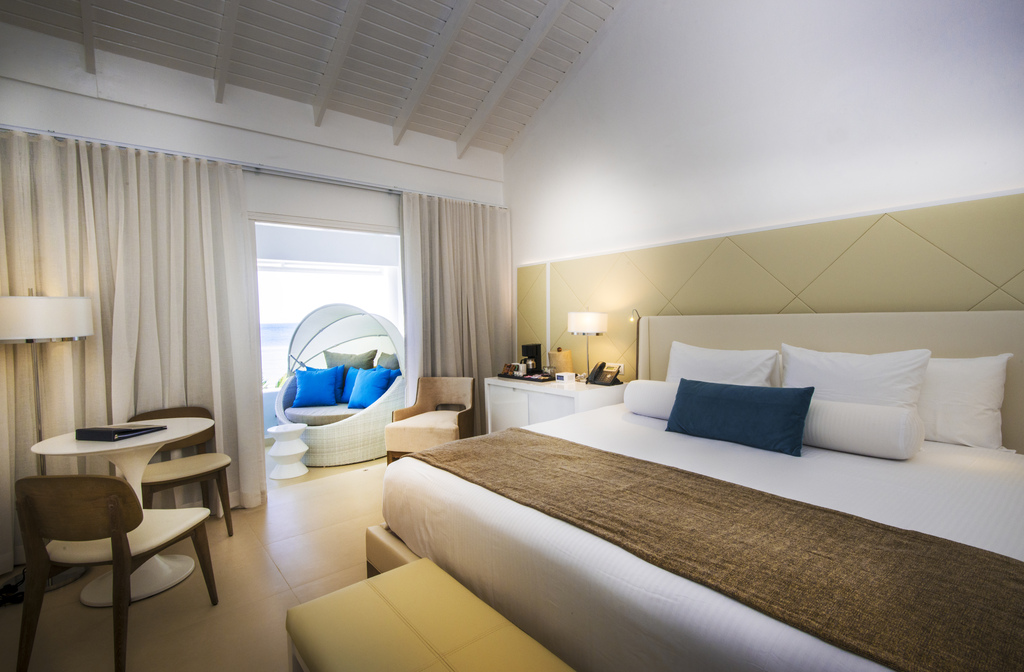 Modern resort bedroom with king bed, sitting area, and curved chair by window overlooking ocean at Azul Beach Resort Negril.