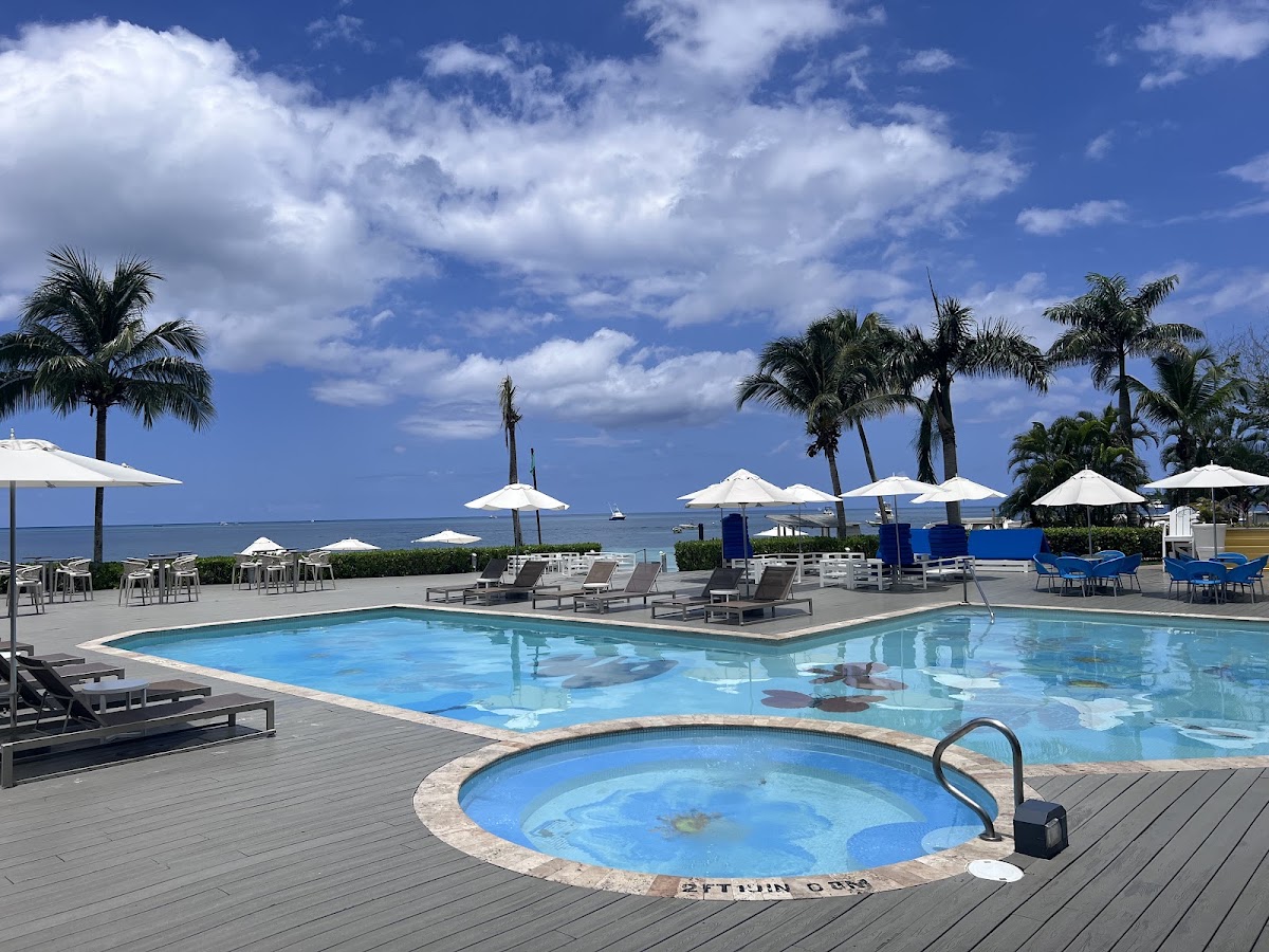 Resort pool deck with hot tub in foreground, main swimming pool beyond, white umbrellas and lounge chairs, palm trees, and calm ocean under blue sky with white clouds.