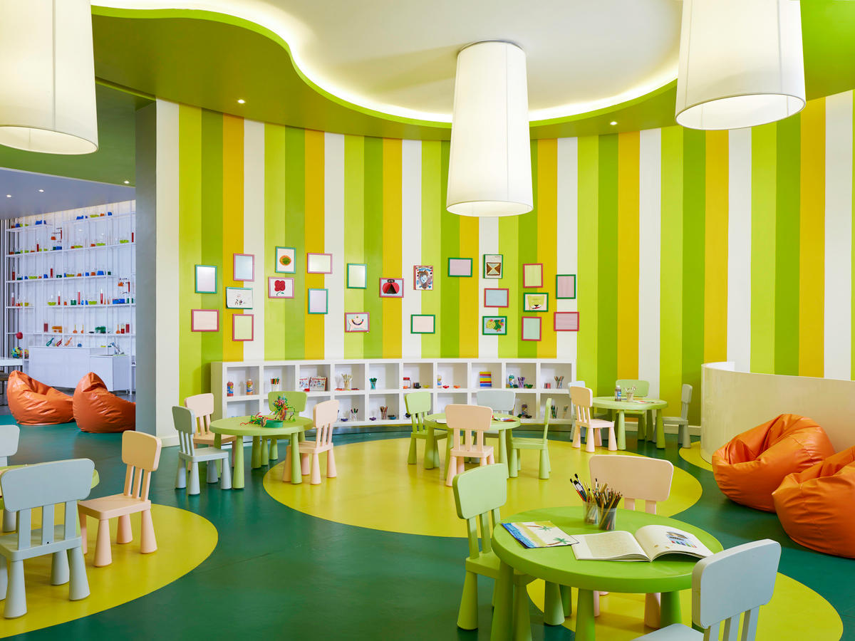 Colorful kids' activity room with lime green and yellow striped walls, small wooden tables and chairs, orange bean bags, and artwork displays at Azul Beach Resort Negril.