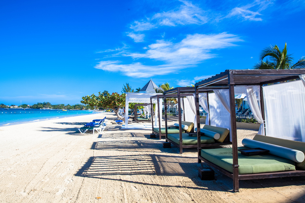 Beachfront cabanas with white curtains and green cushions line the sand at Azul Beach Resort Negril, with turquoise water and palm trees beyond.