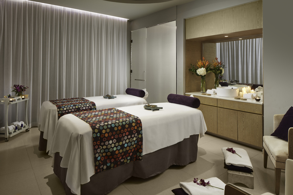 Spa treatment room with two massage beds draped in white linens, patterned runners, and dark bolsters, with a wood vanity holding fresh flowers and candles across from floor-to-ceiling curtains.