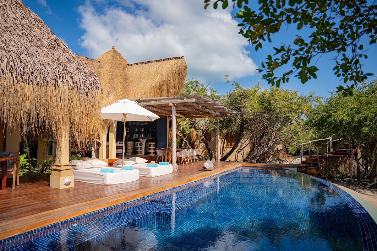 Blue-tiled pool with wooden deck facing a thatched-roof villa with white lounge beds and pergola at Azura Benguerra Island.