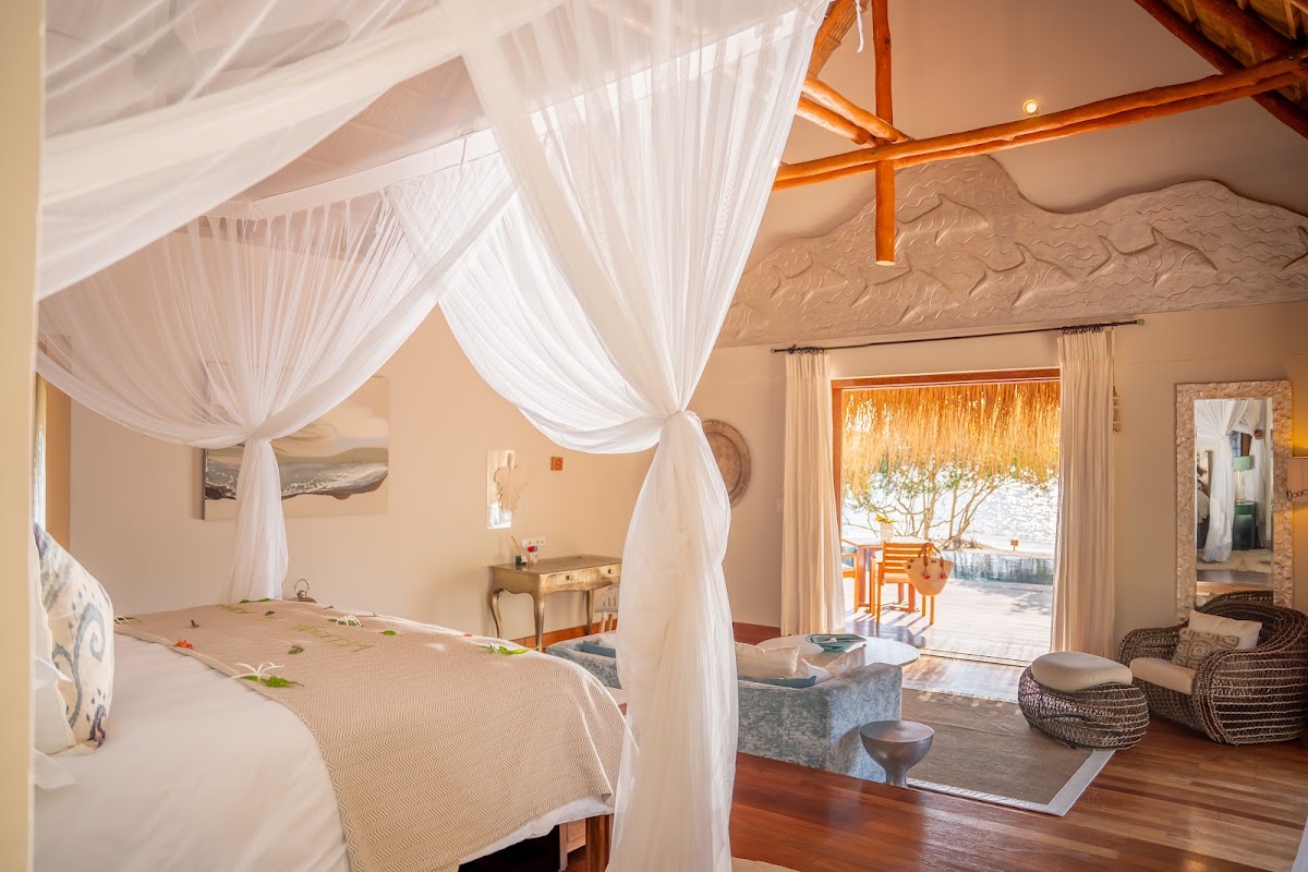 Four-poster bed with white sheer curtains in a spacious resort suite opening to an ocean view terrace at Azura Benguerra Island.