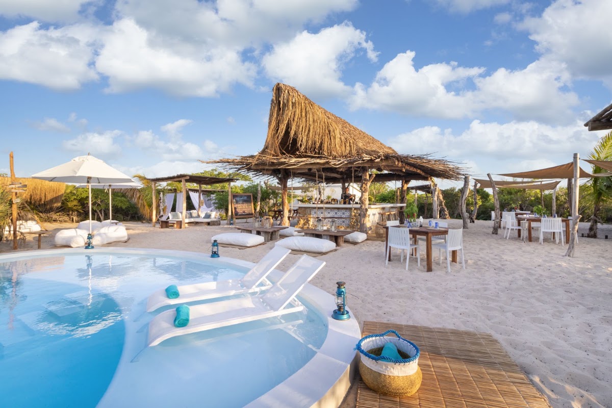 Beach club at Azura Benguerra Island with loungers by a pool, thatched-roof shelters, dining areas, and sand beneath a blue sky.