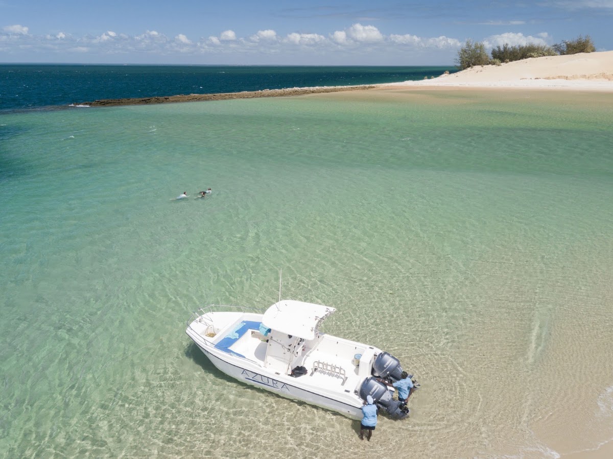 White motorboat moored in shallow turquoise water off a sandy beach at Azura Benguerra Island, with swimmers visible in the distance.