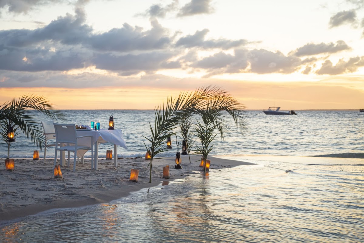 Beach dinner setup at sunset with palm fronds, lanterns, and a table on the sand at Azura Benguerra Island.