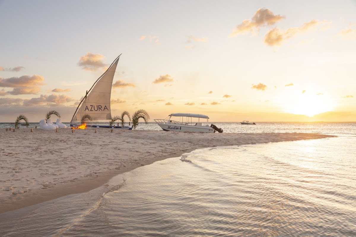 Dhow sailboat with Azura branding and motorboats on sandy beach at sunset with gentle waves and palm decorations.