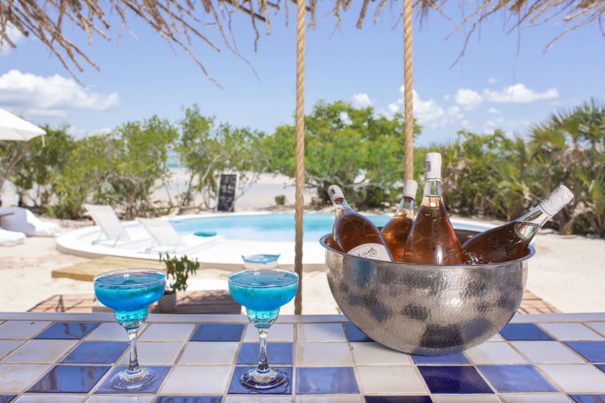 Two blue cocktails and a metal ice bucket with champagne bottles sit on a tiled table under rope swings, overlooking a pool and palm trees at Azura Benguerra Island.